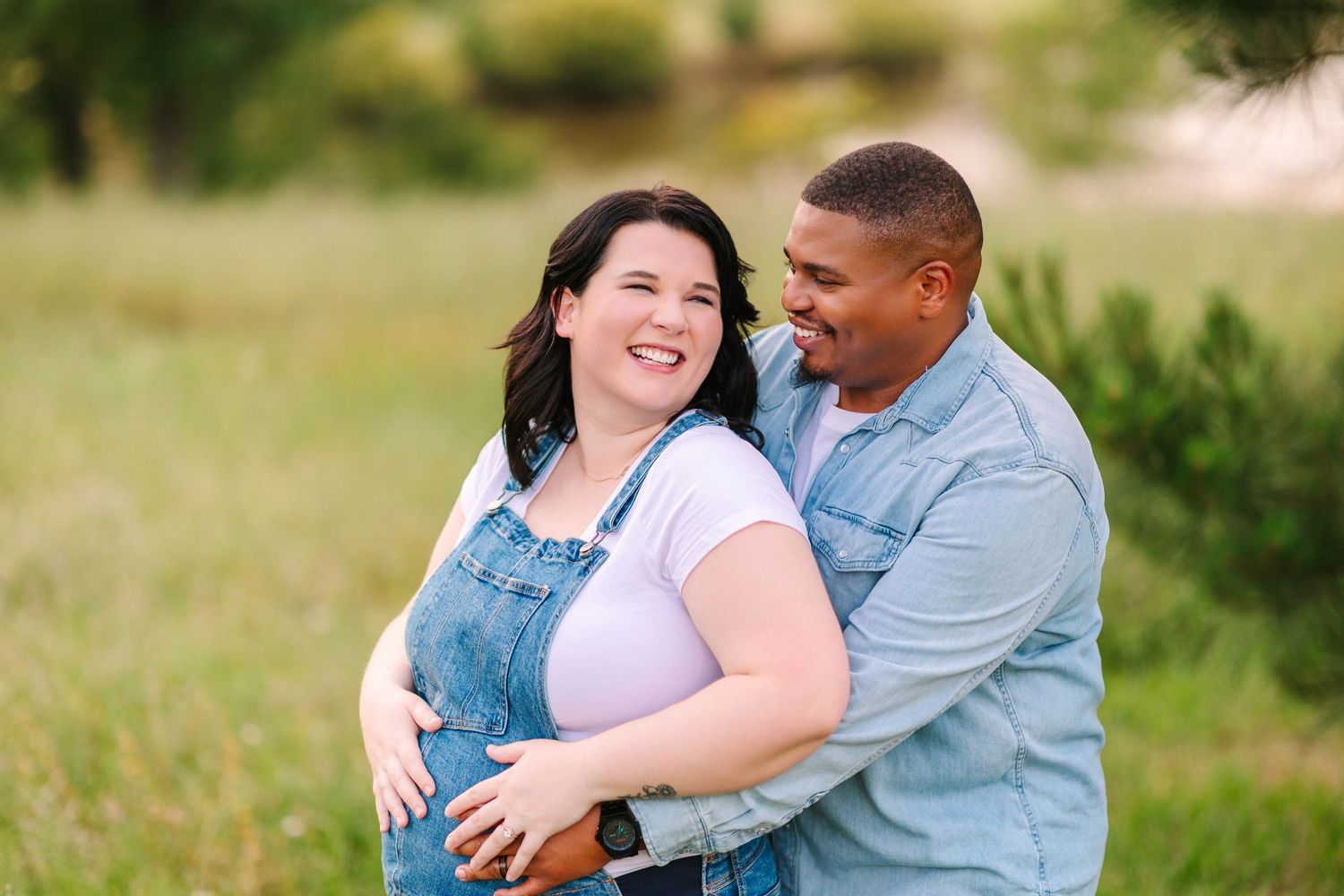A couple shares a joyful moment together while embracing in a sunlit grassy field during a maternity photo session.
