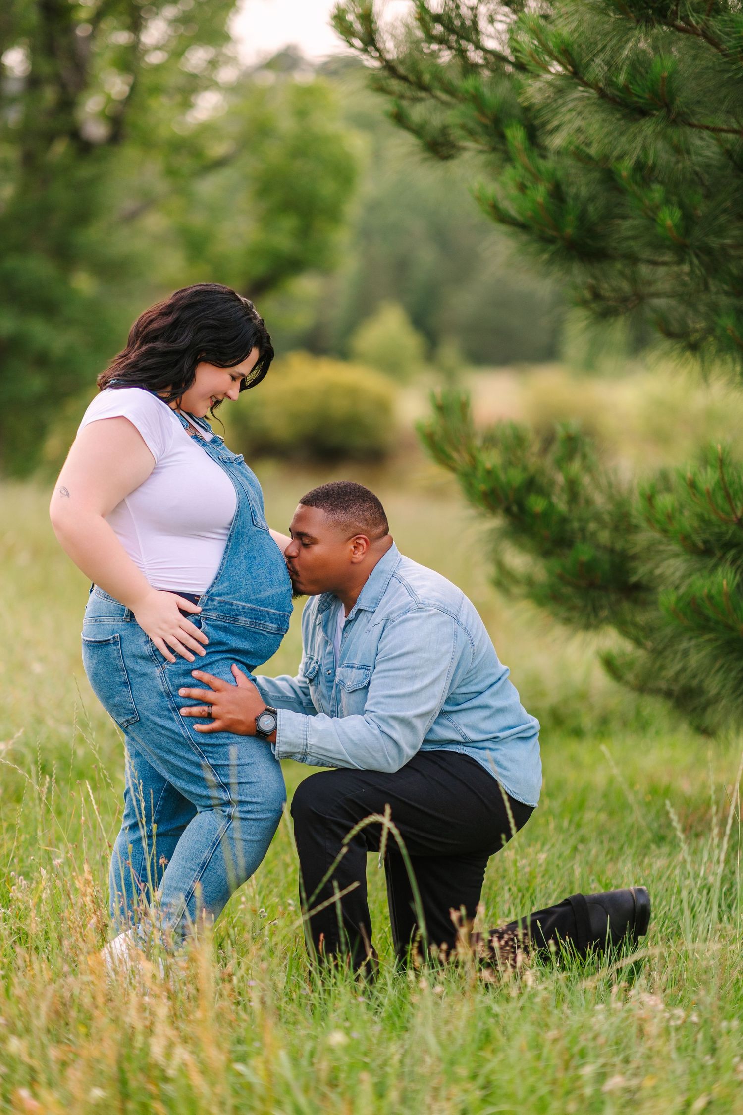 Tender outdoor maternity moment captured as couple poses near evergreen trees in a natural setting.