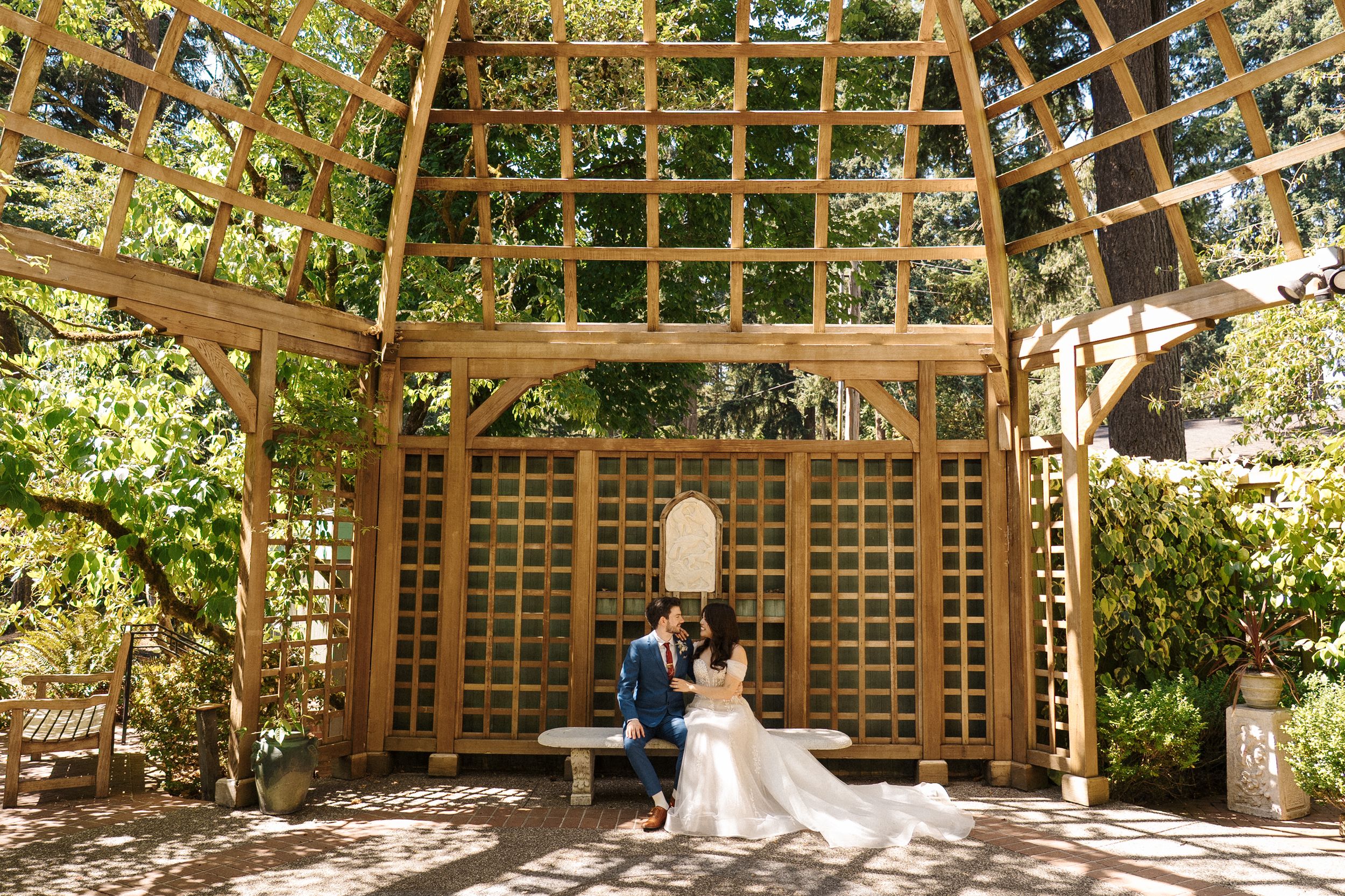 A couple sits together in a sunlit wooden pergola surrounded by climbing vines and garden greenery.
