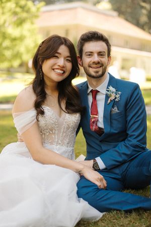 A couple in wedding attire sit together and smile on a sunny lawn with natural lighting.