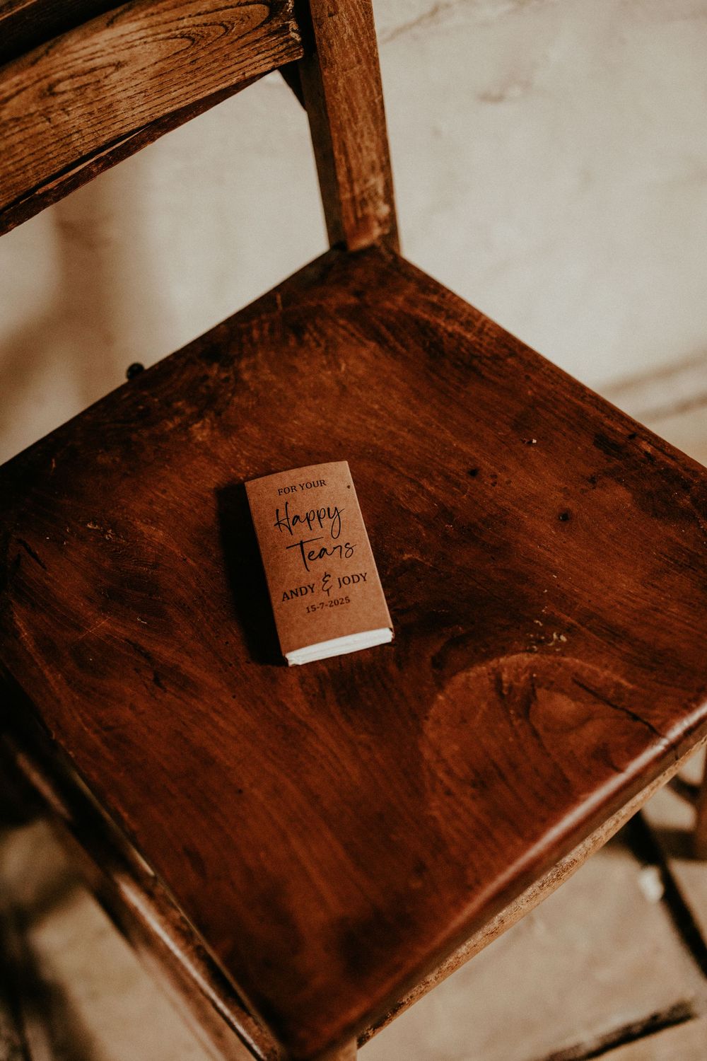 A small brown book titled "Happy Tears" rests on a rustic wooden chair seat in moody lighting.