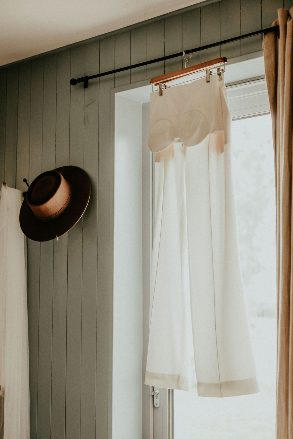 Simple white curtains with wood trim hanging against sage green vertical paneled walls in a rustic interior.