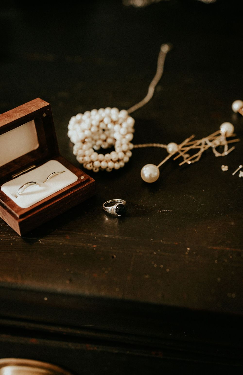 Wedding rings in a wooden box alongside pearl jewelry on a dark wooden surface in moody lighting.