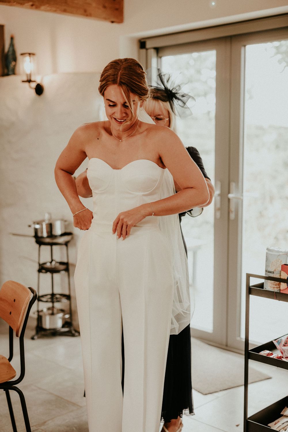 Wedding dress fitting scene showing final adjustments being made to a classic white gown in a bright sunlit room.