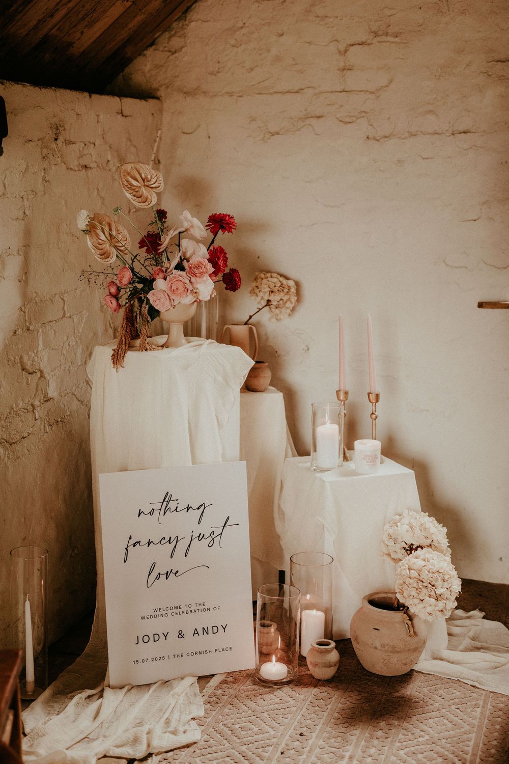 A series of ivory-colored wedding decor items with handwritten font and floral arrangements set against a rustic stone wall.