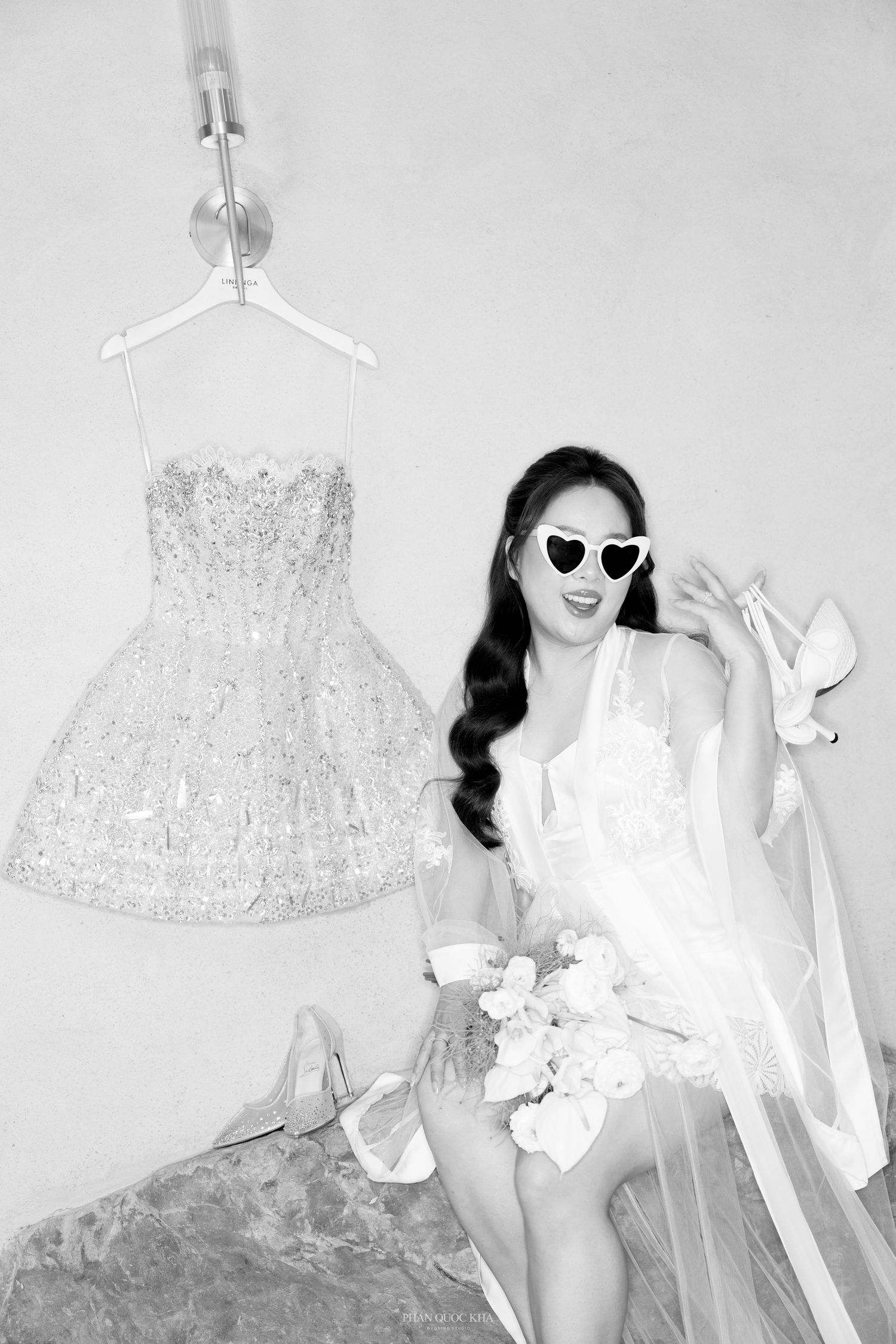 Black and white photo of a bride in heart sunglasses posing beside a sparkling mini wedding dress.