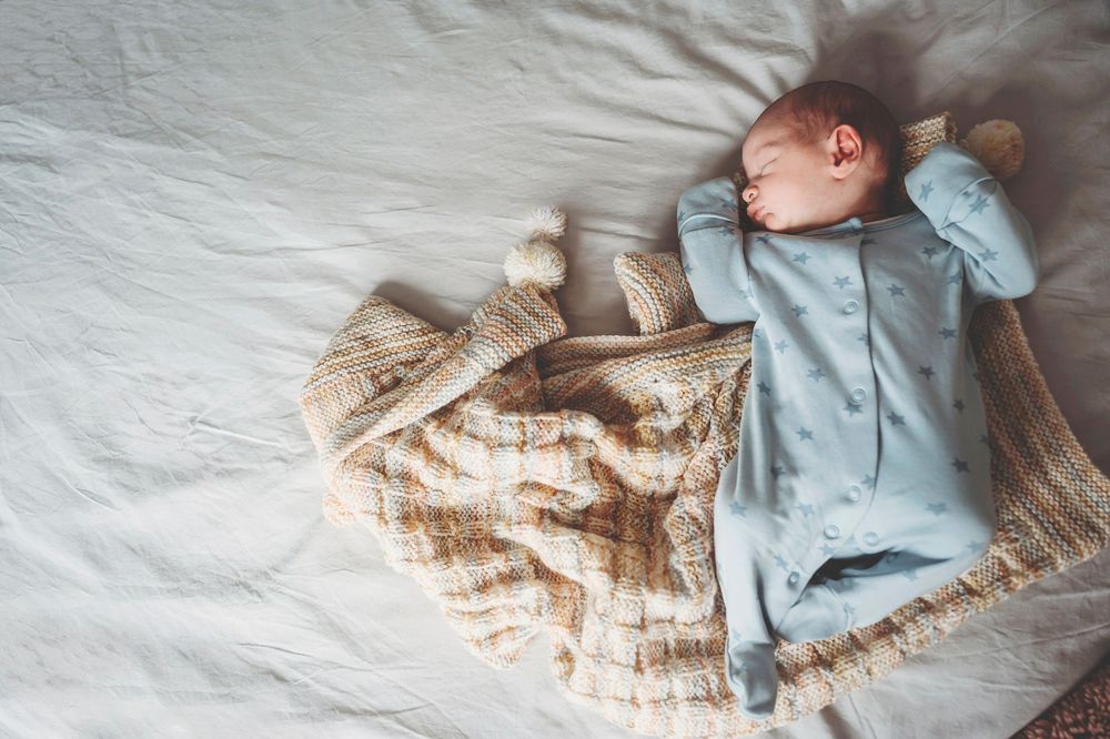 A sleeping baby wearing a light blue outfit lays peacefully on white and beige bedding under soft lighting.
