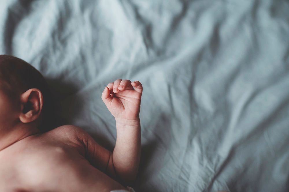 Baby wearing white fabric on bed in sleeping or napping position with arms stretched upwards.