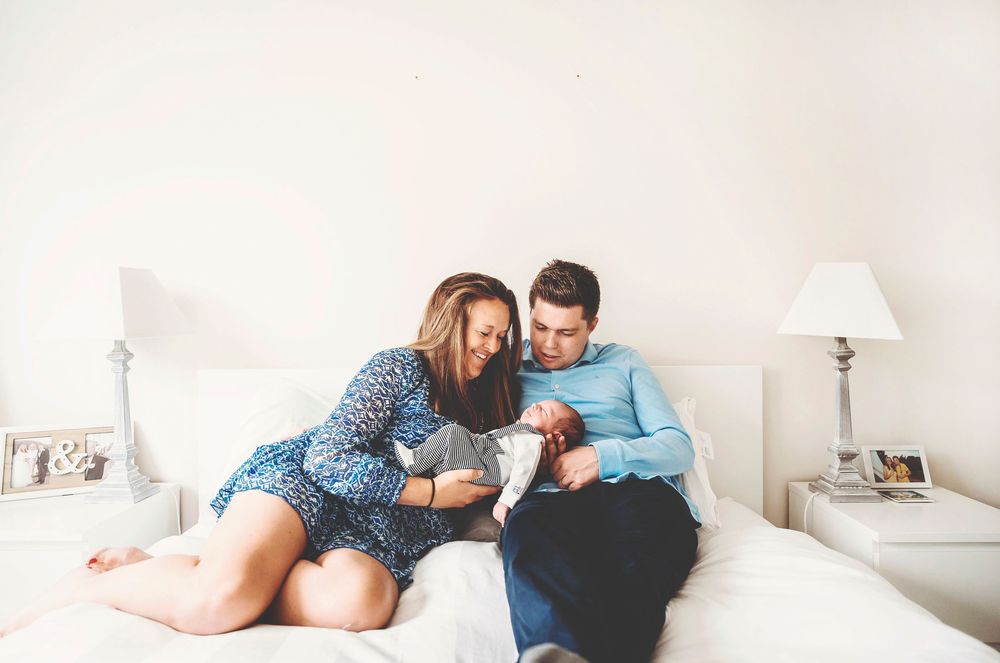 Couple relaxing together on white bed looking at small baby in blue shirt's arms.