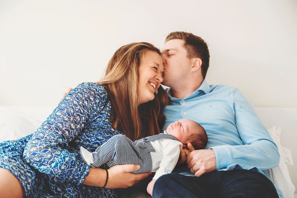 Parents share joyful moment on bed while holding their newborn baby in grey blanket.