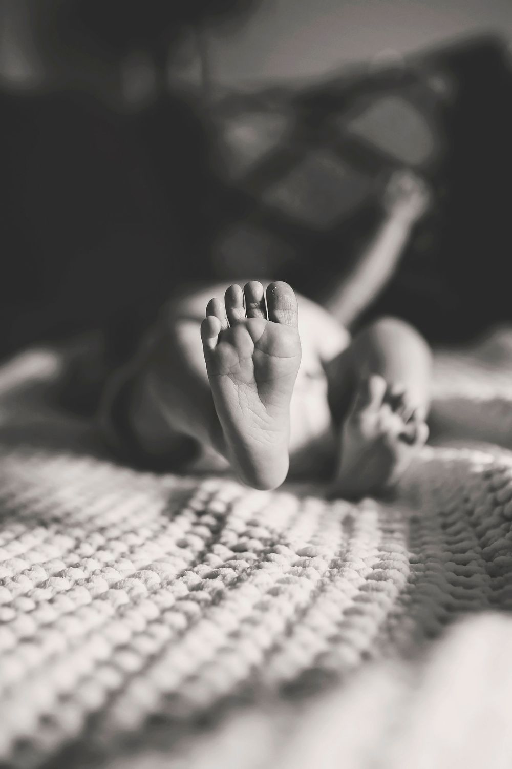 Black and white close-up photo shows a small baby foot against a soft textured surface creating visual contrast.