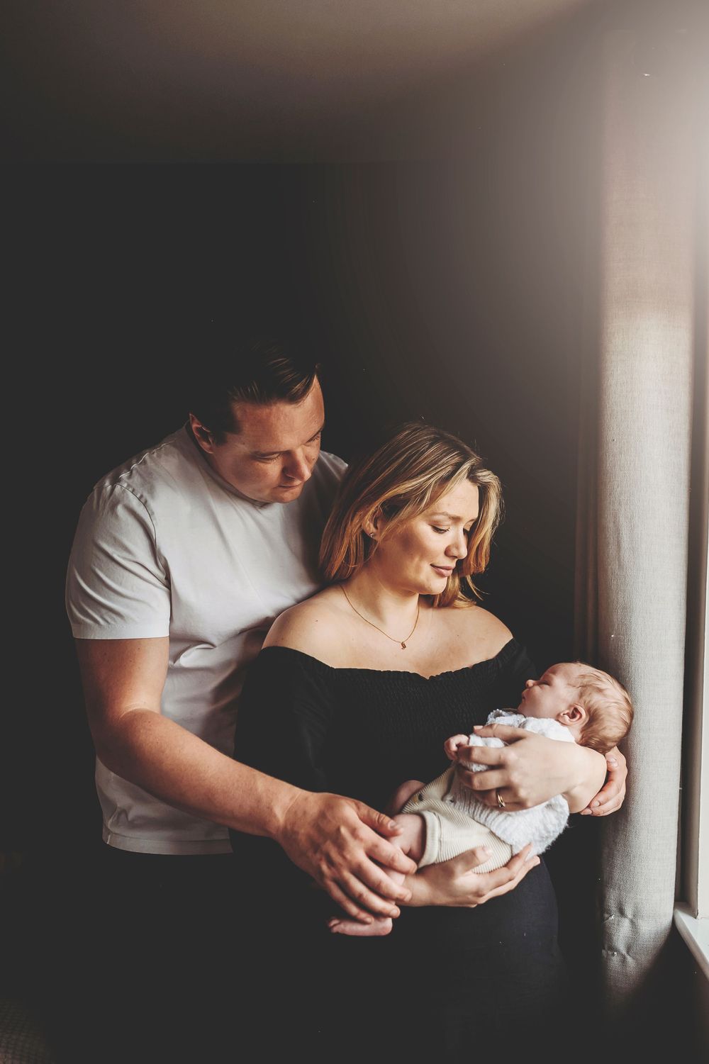Tender moment between parent and newborn baby shown in a series of moody, intimate portraits by a window.