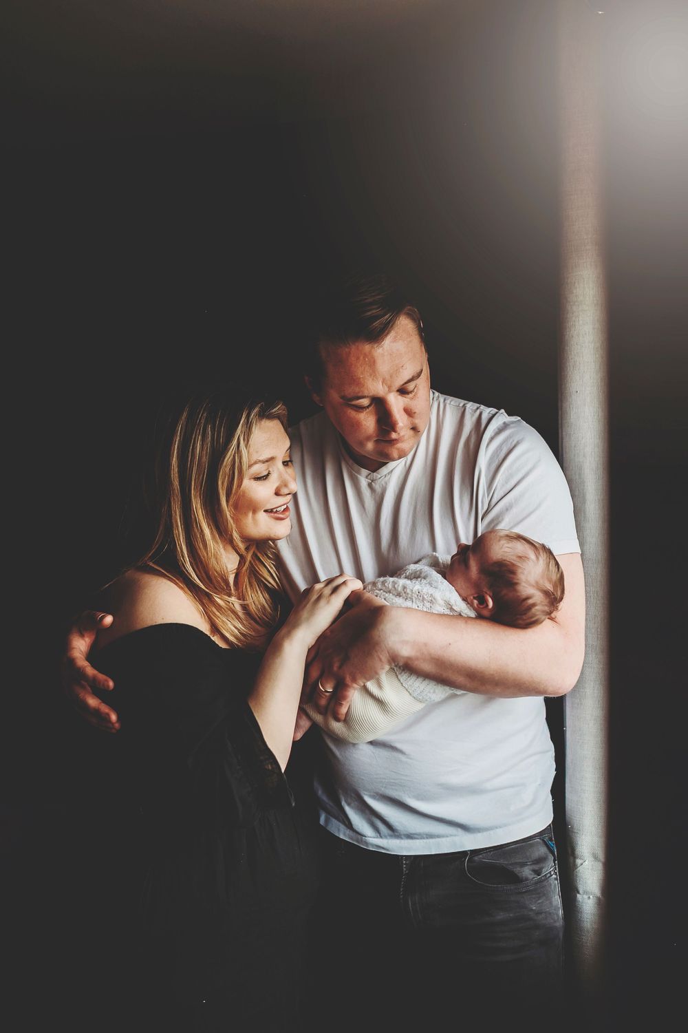A couple in a moody, dark setting embrace tenderly against a light-filled window creating an intimate silhouetted moment.
