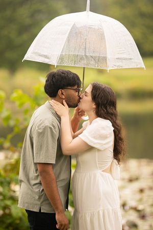 A couple shares an intimate moment under a clear umbrella by a scenic pond.