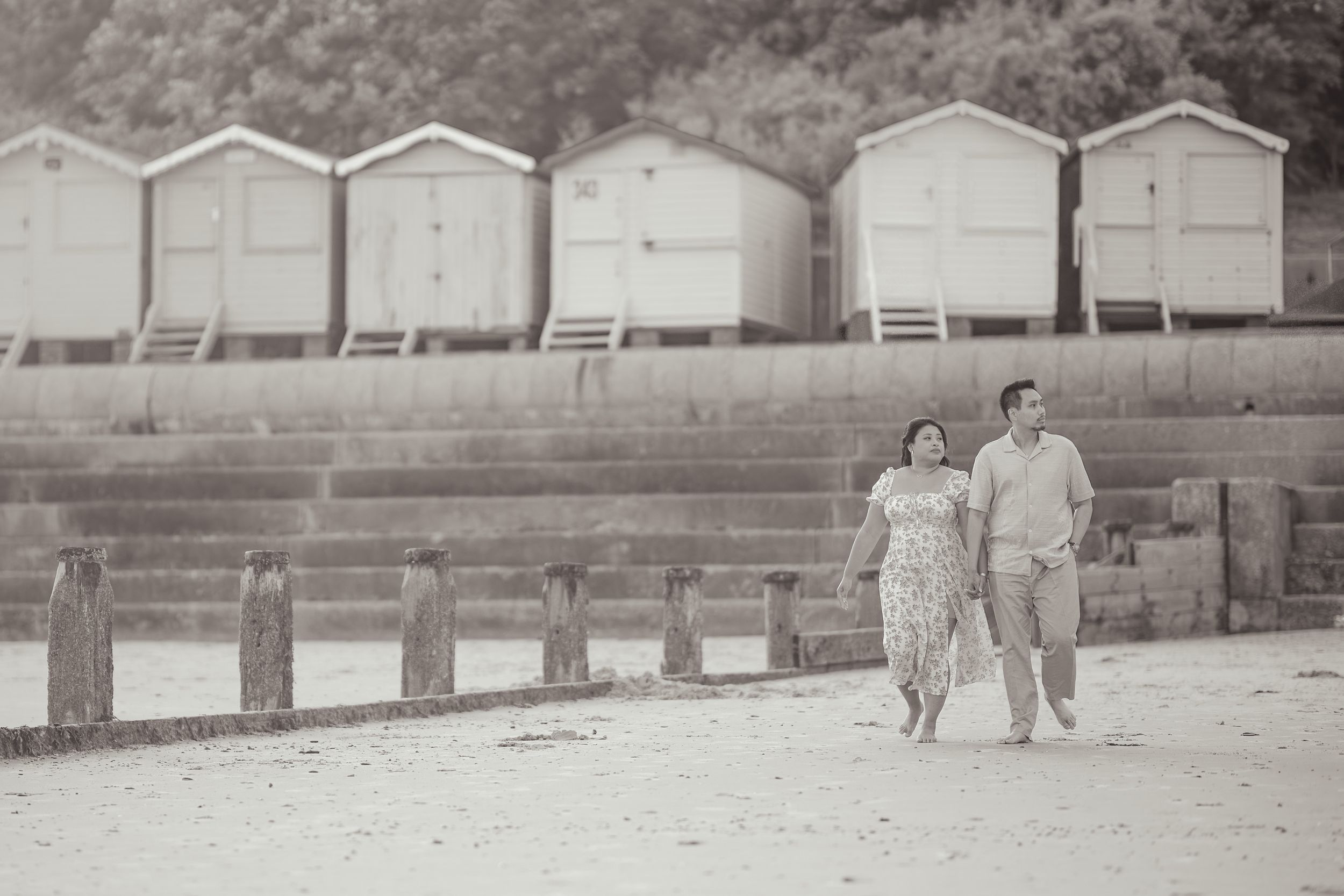 A couple strolls past vintage beach huts in a black and white photograph.
