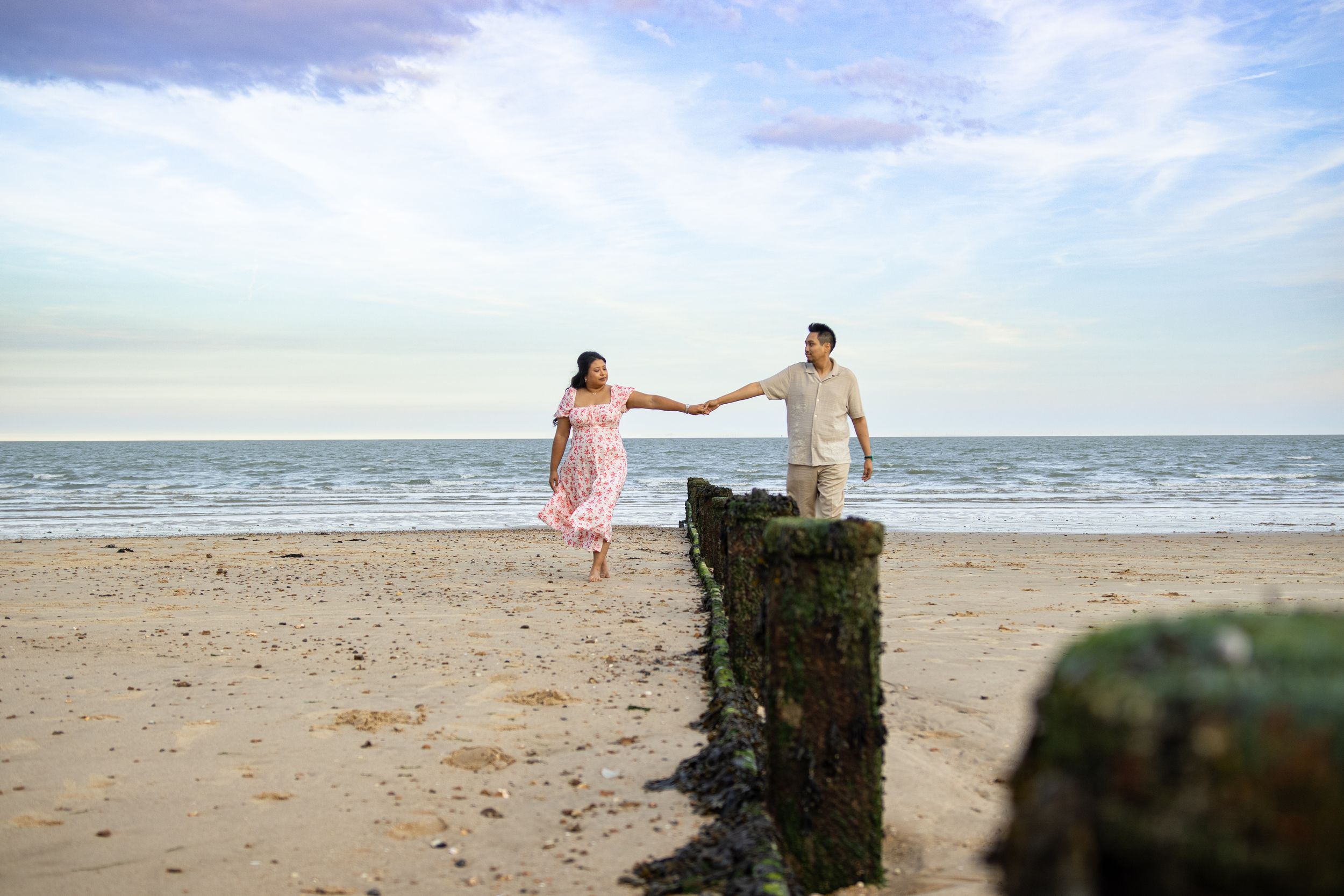 A couple walks hand in hand along a serene beach with wooden posts in the foreground.