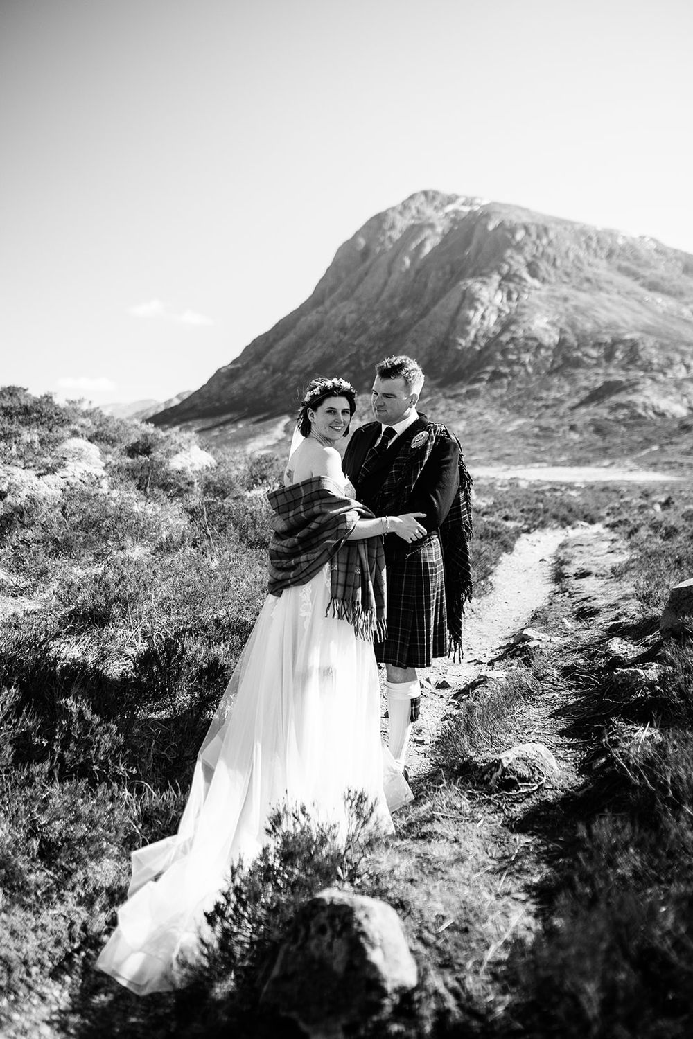 A couple embrace outdoors in traditional Scottish Highland dress against a dramatic mountain landscape in black and white.