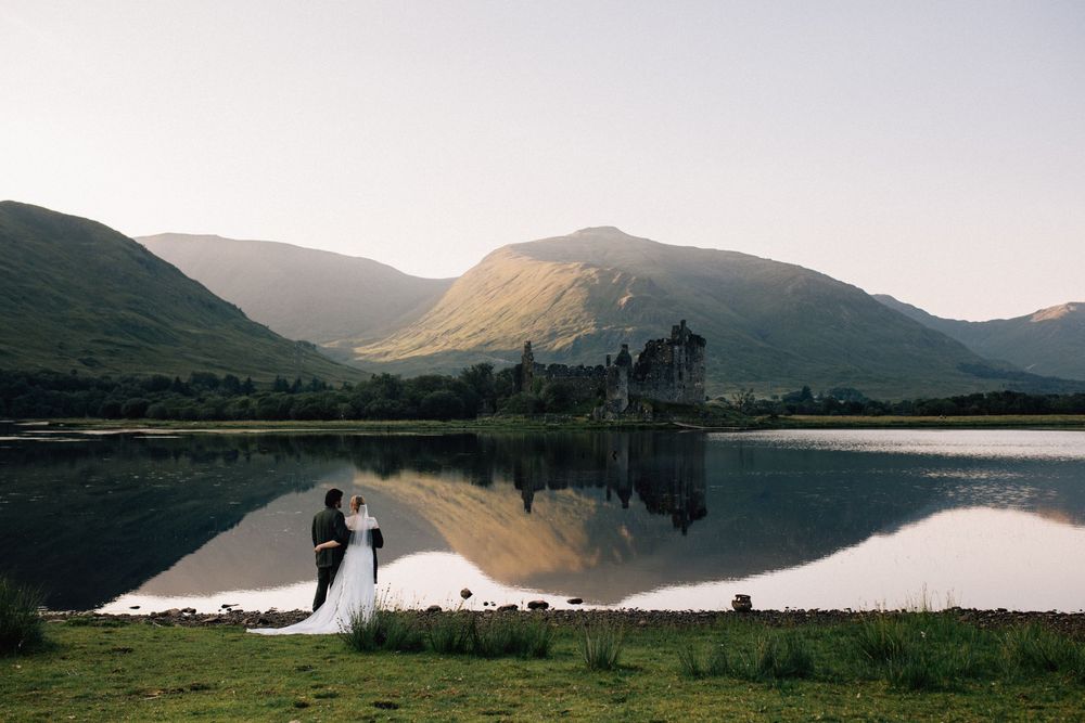 Bride and groom stand by a reflective loch with a Scottish castle and misty green mountains at sunset.