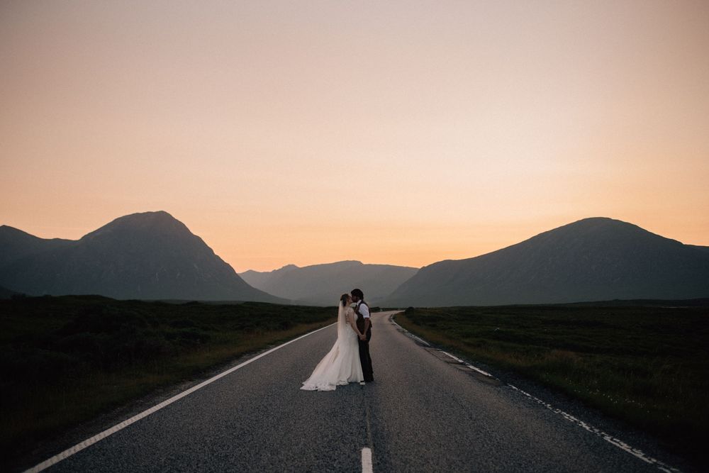 Bride and groom kissing on a scenic road at sunset with mountain silhouettes in the background.