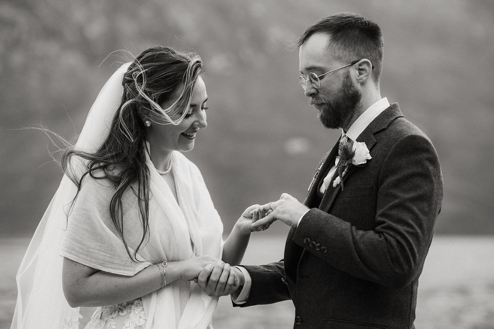 Black and white photo of a groom placing a wedding ring on his bride's finger during an outdoor ceremony.