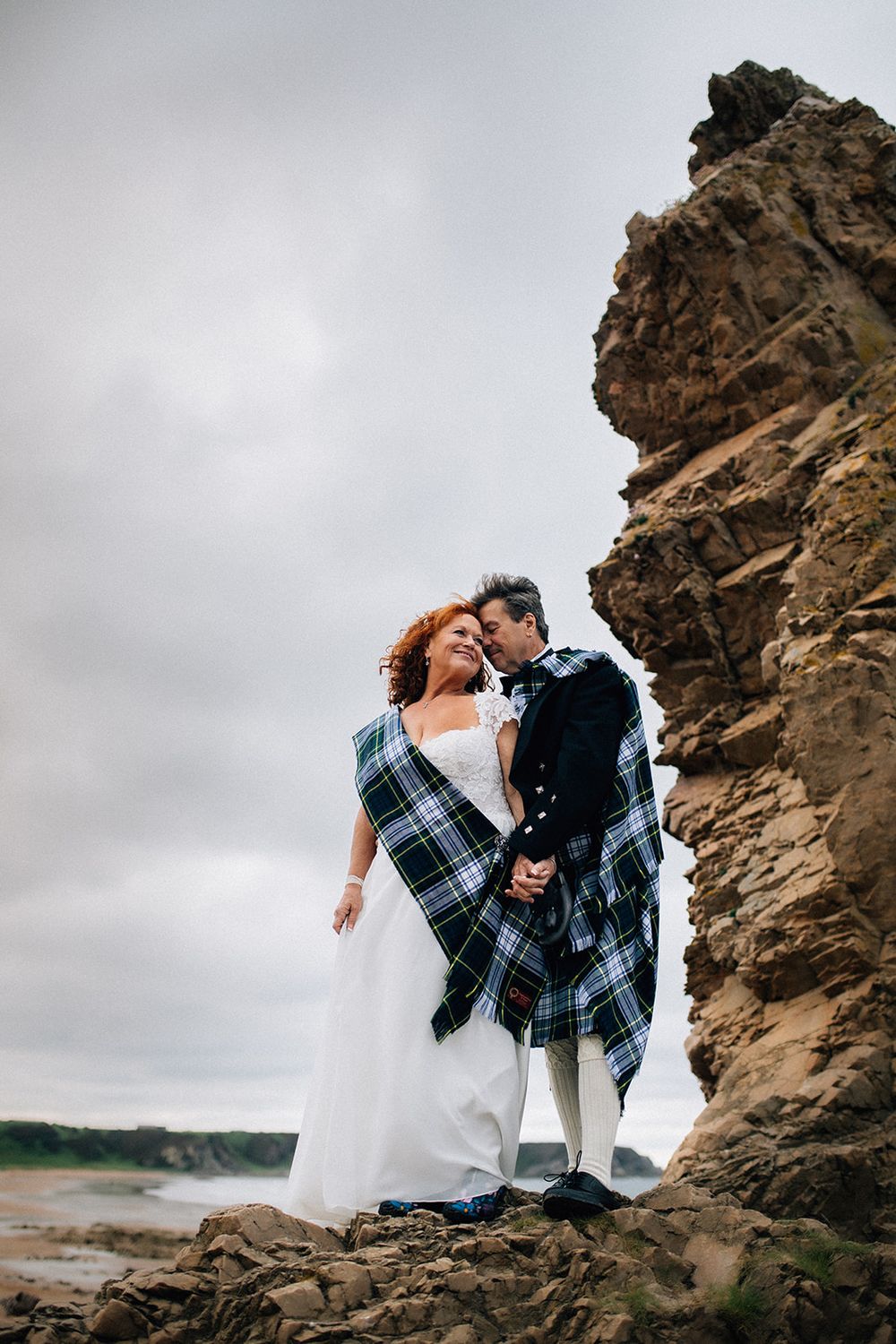 Bride and groom in Scottish tartan embrace on rocky coastal cliffs on their wedding day.