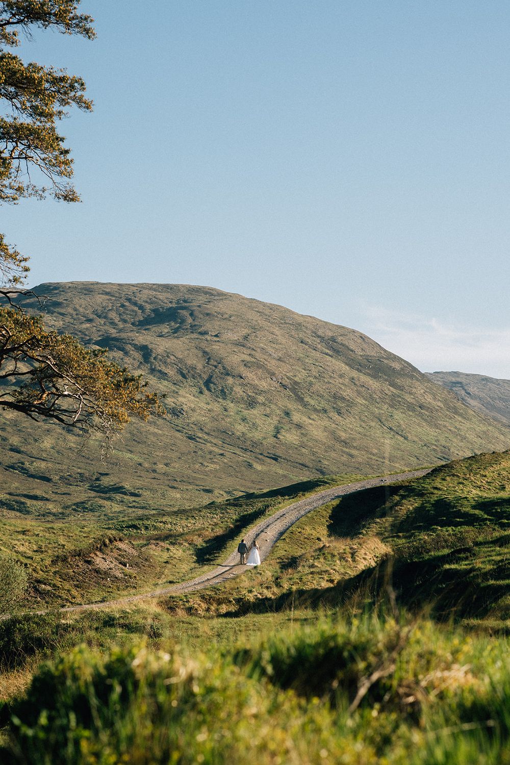 A couple walks along a winding road through lush green Scottish Highlands with a majestic mountain backdrop.