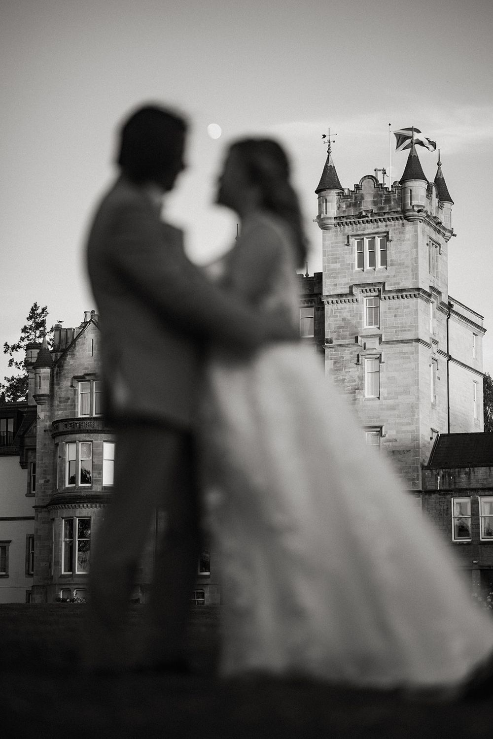 Black and white wedding photo of couple embracing in front of a grand castle at dusk.