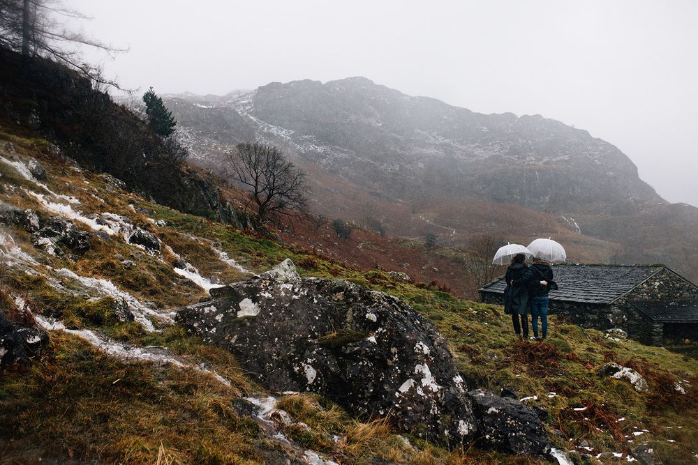 Two people with umbrellas walk along a snowy mountain trail in foggy weather near a stone building in Scotland.
