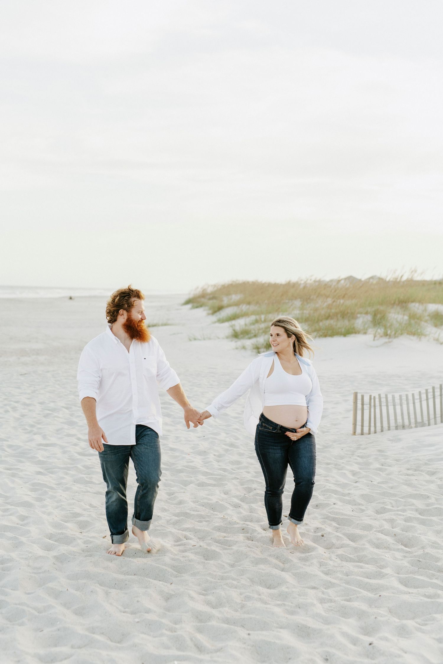 A couple playfully running and interacting on a serene sandy beach with grass-covered dunes in the background.