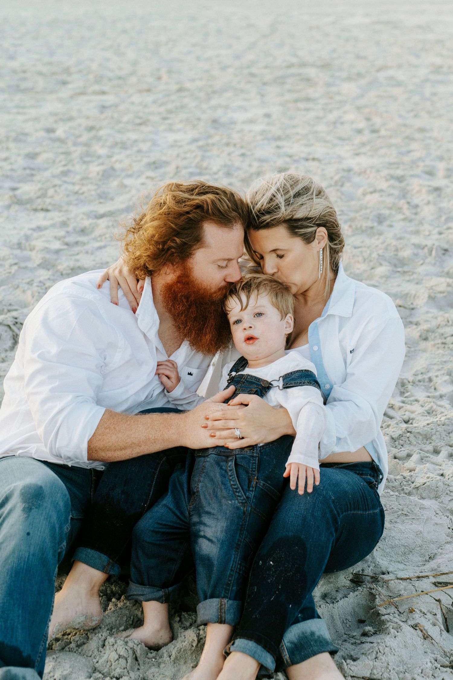 A family wearing white shirts and jeans shares intimate moments while sitting together at a beach.