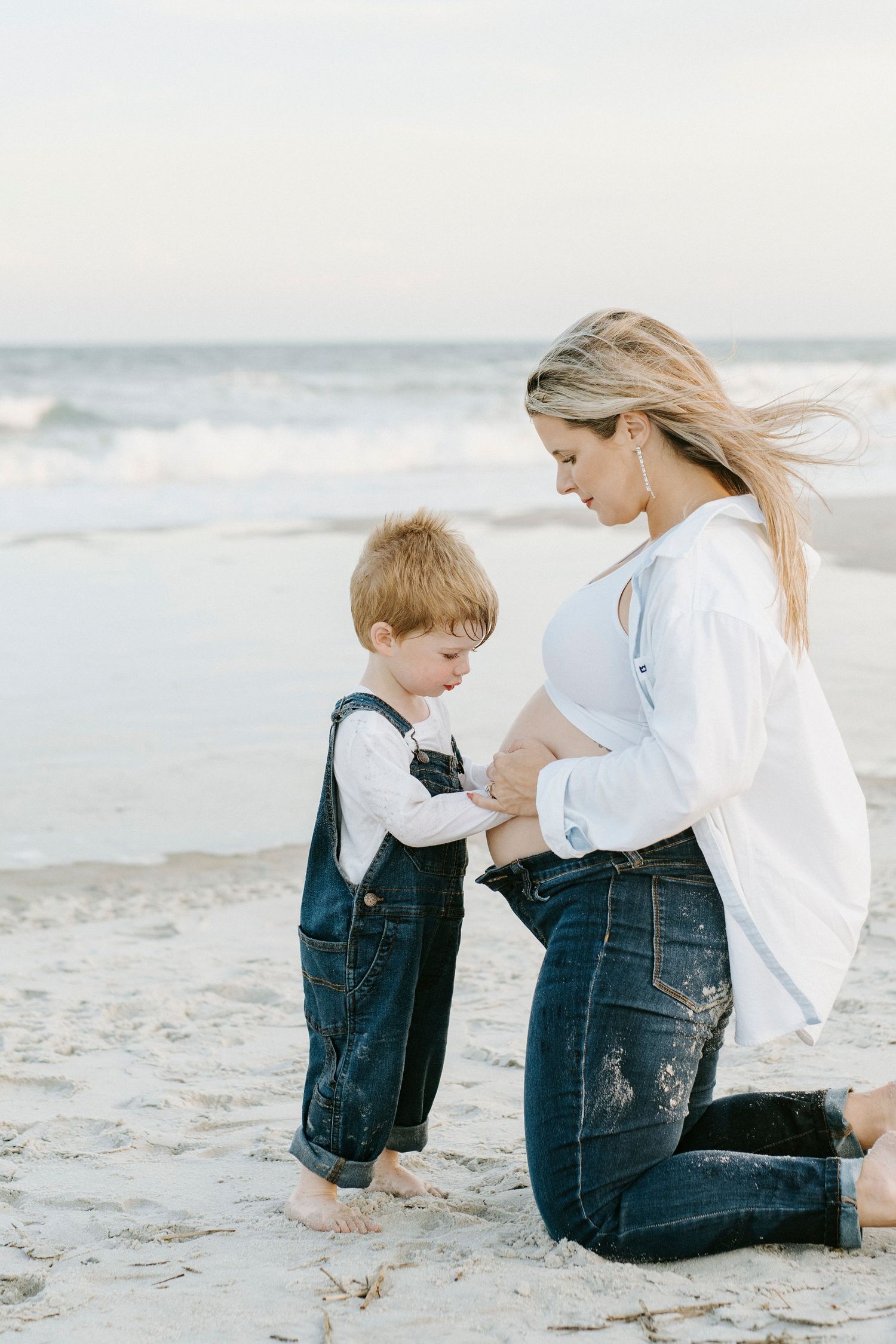 Mother and young child share tender moment while kneeling together on sandy beach at sunset.