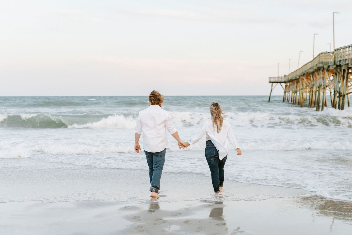 A couple walks hand in hand along a misty beach shoreline with waves crashing near a wooden pier.