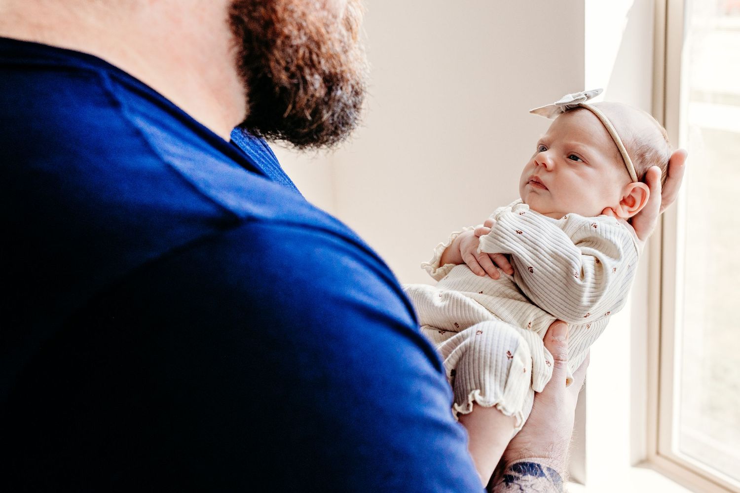 Parent wearing blue shirt holds and cradles newborn baby wearing white outfit next to window in natural light.
