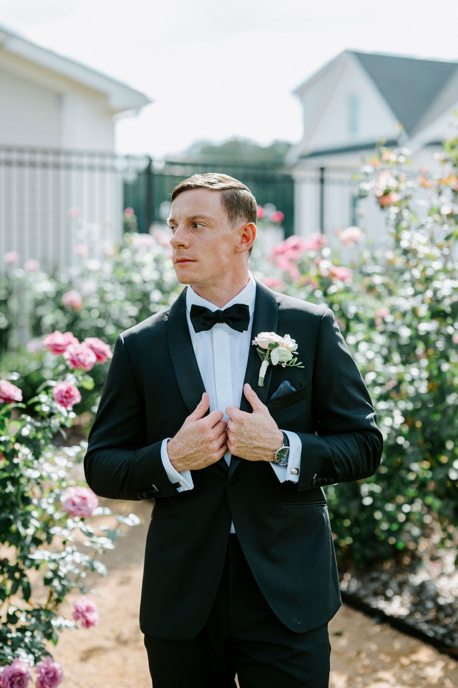Man in black tuxedo and bowtie adjusts his jacket in a garden with blooming roses.