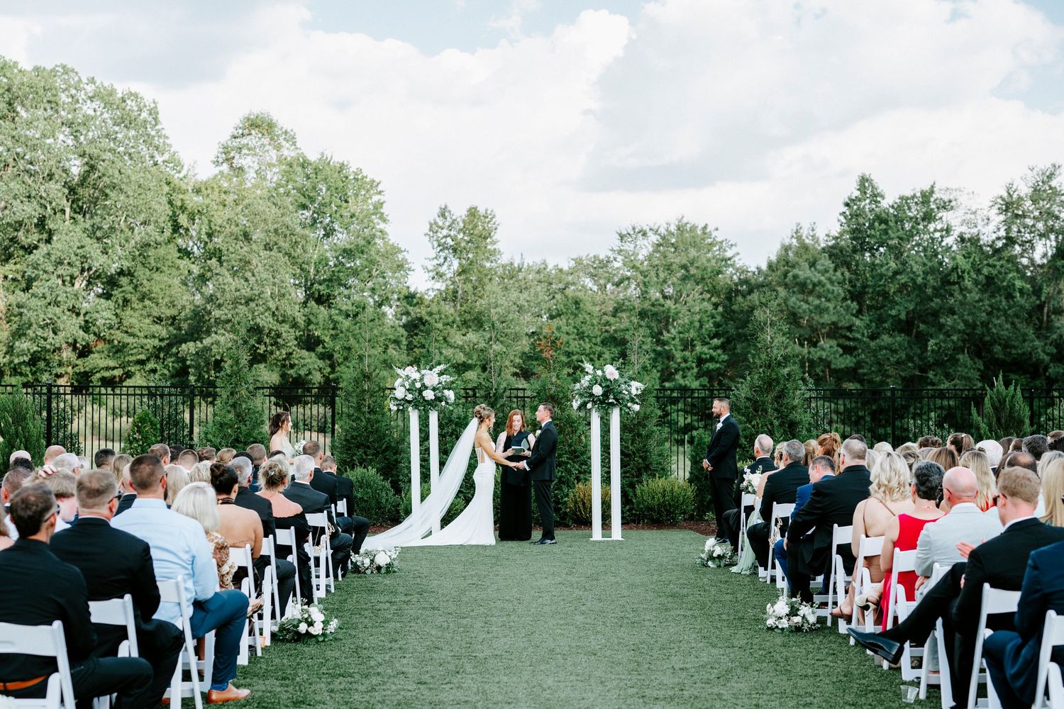 Outdoor wedding ceremony setup with white chairs and arch on a green lawn surrounded by trees under cloudy skies.
