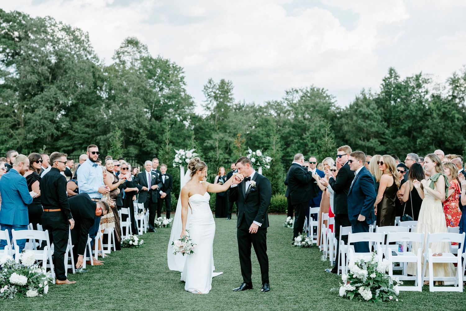 Wedding guests stand during outdoor ceremony as couple celebrates at altar with white chairs lining the grass aisle.