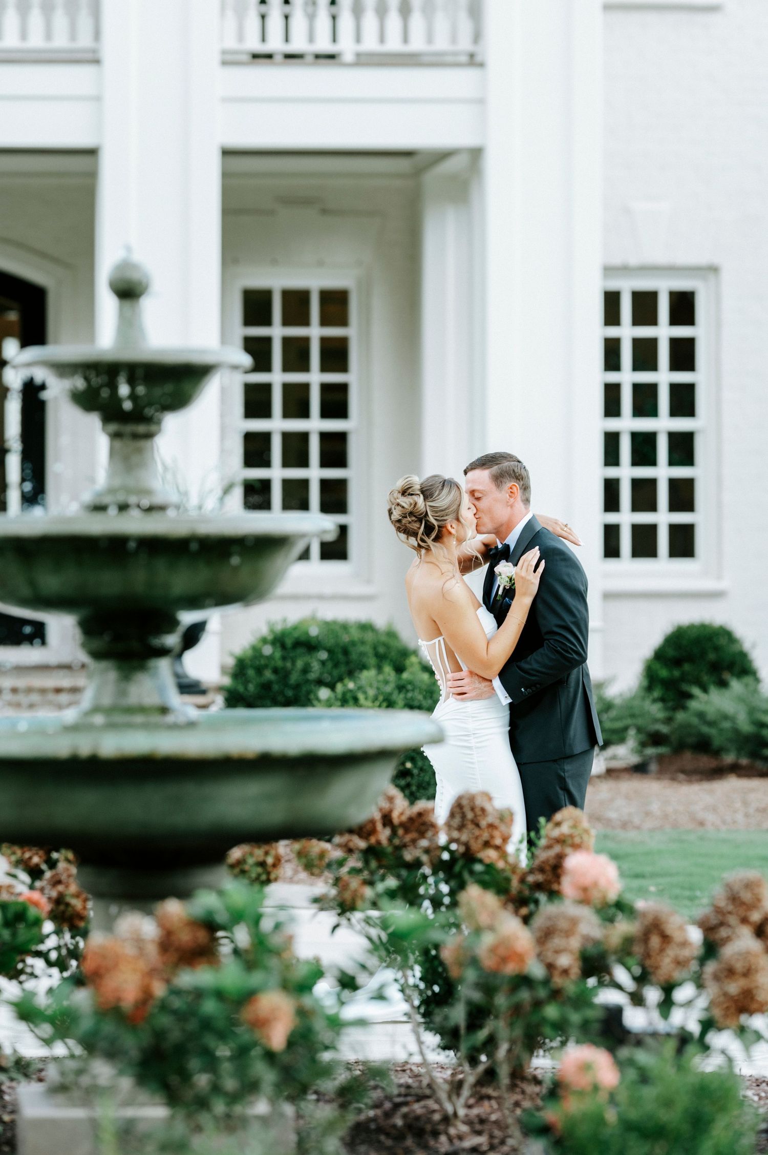 A romantic moment between newlyweds by an elegant fountain outside a grand white mansion with colonial architecture.