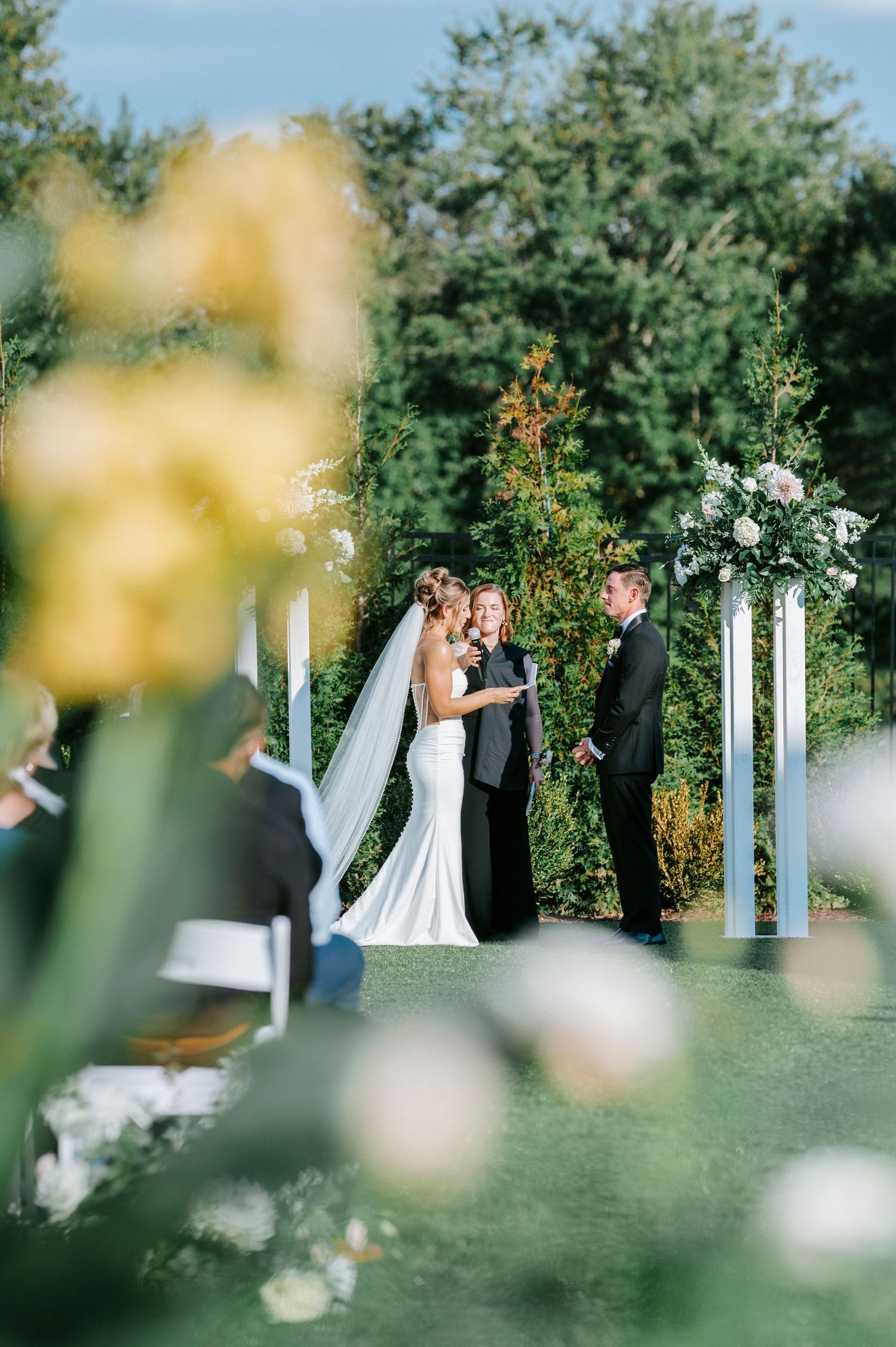 Outdoor wedding ceremony setup in garden with white columns and floral decorations against lush greenery.
