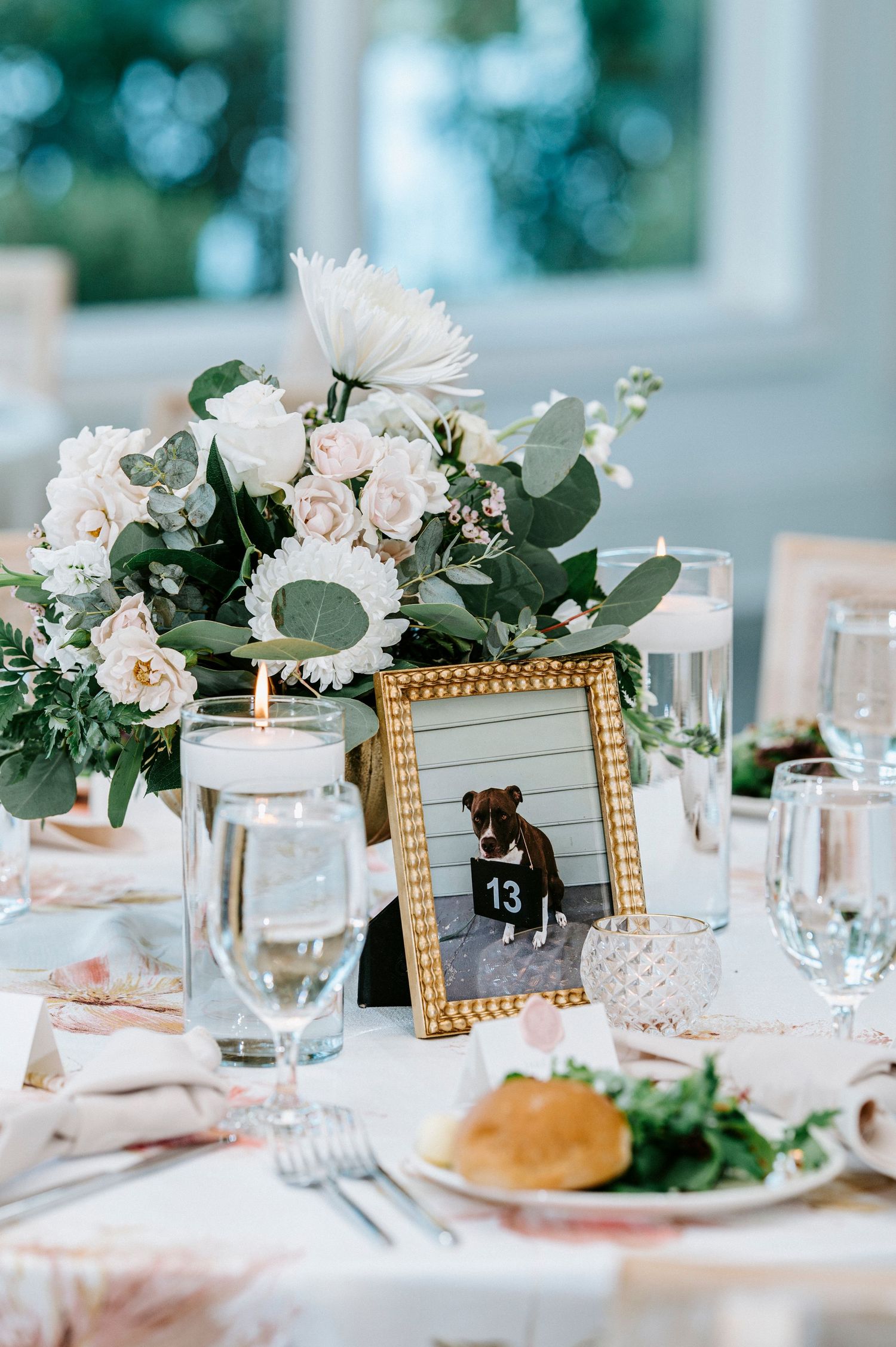 Wedding reception table with gold frames, white flowers, and place settings against a soft blurred background.