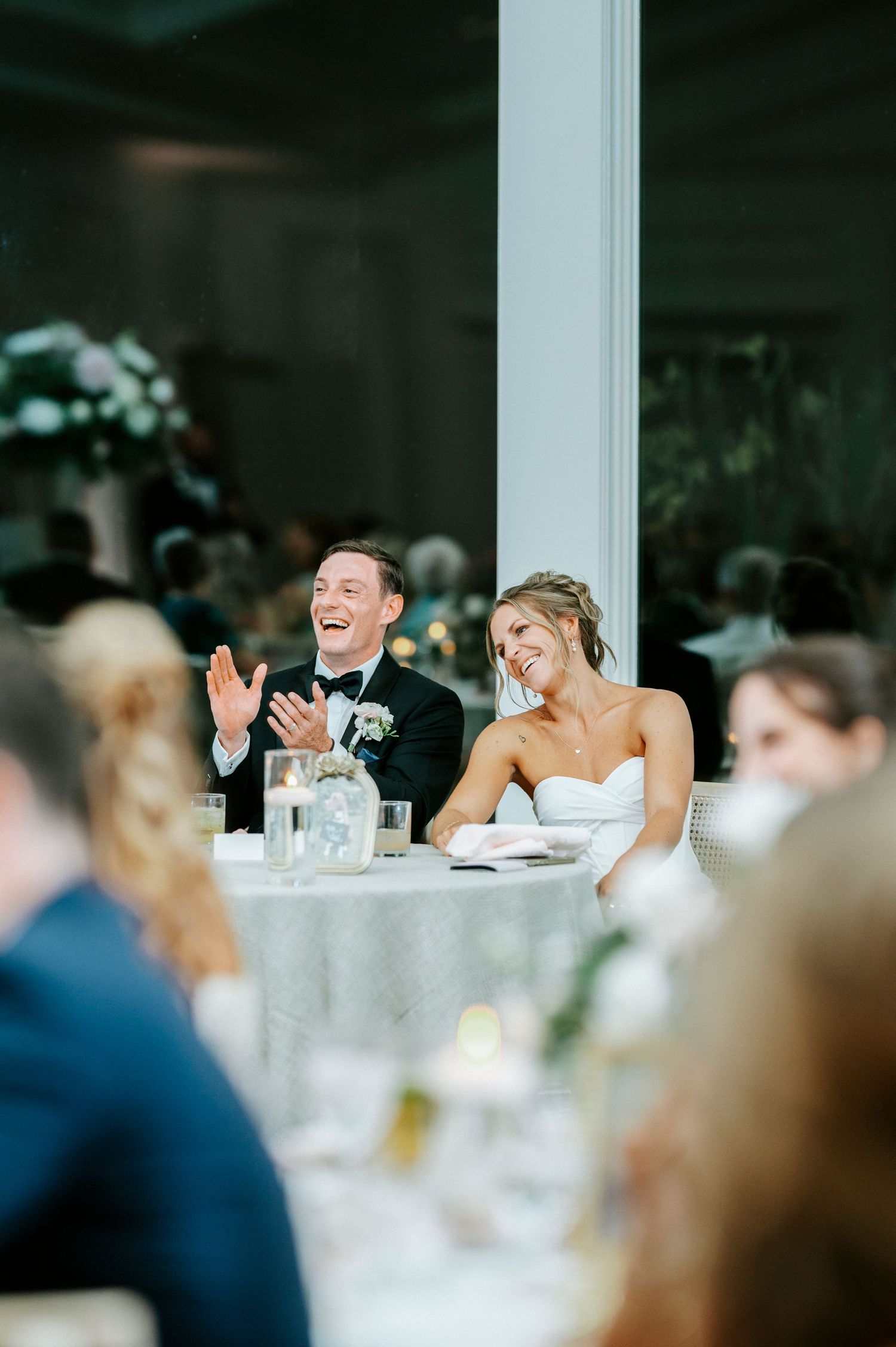 Wedding reception dinner guests seated at elegant white tables with candlelight ambiance indoors.