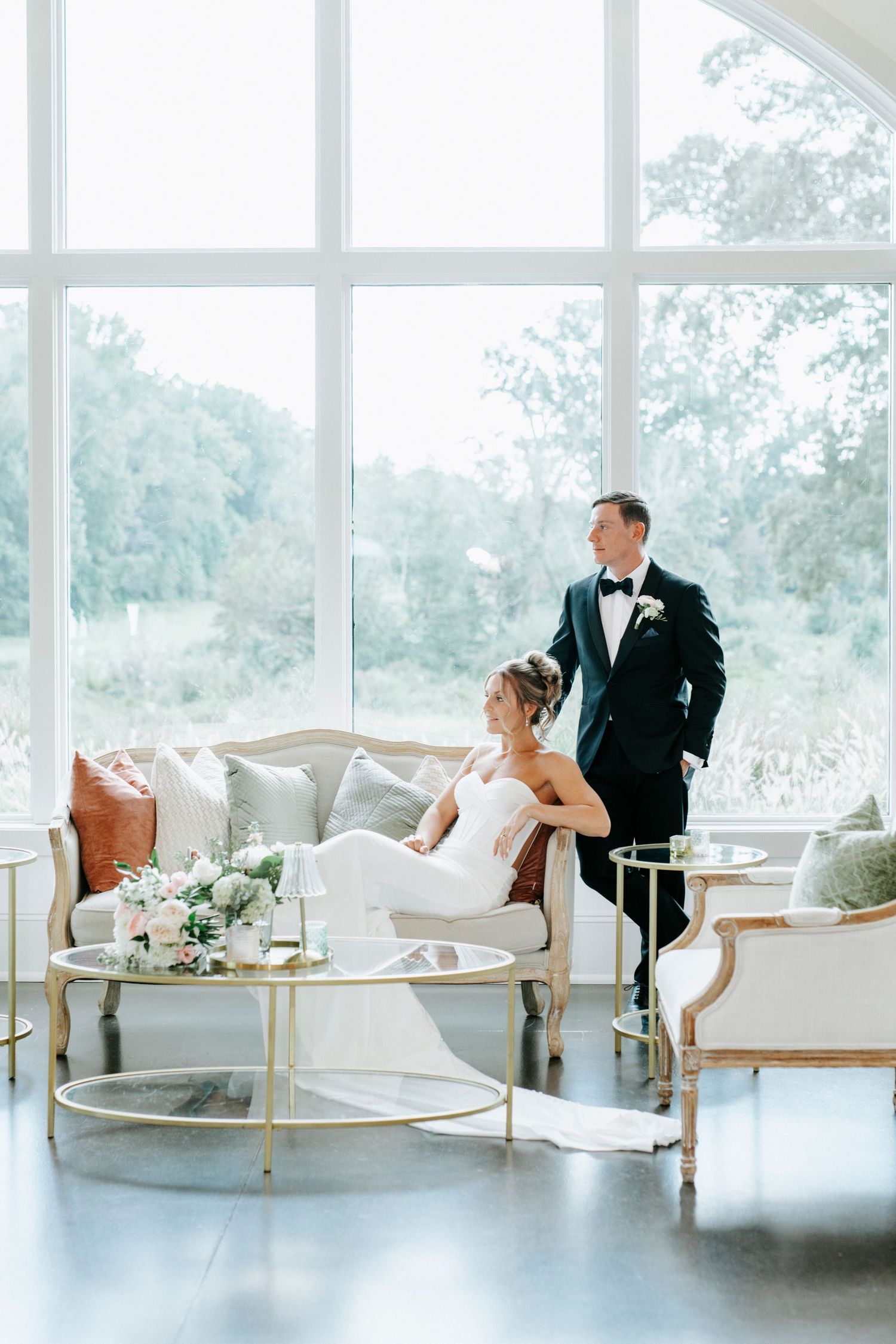 Elegant wedding couple poses in bright sunlit room with modern furniture and floor to ceiling windows.