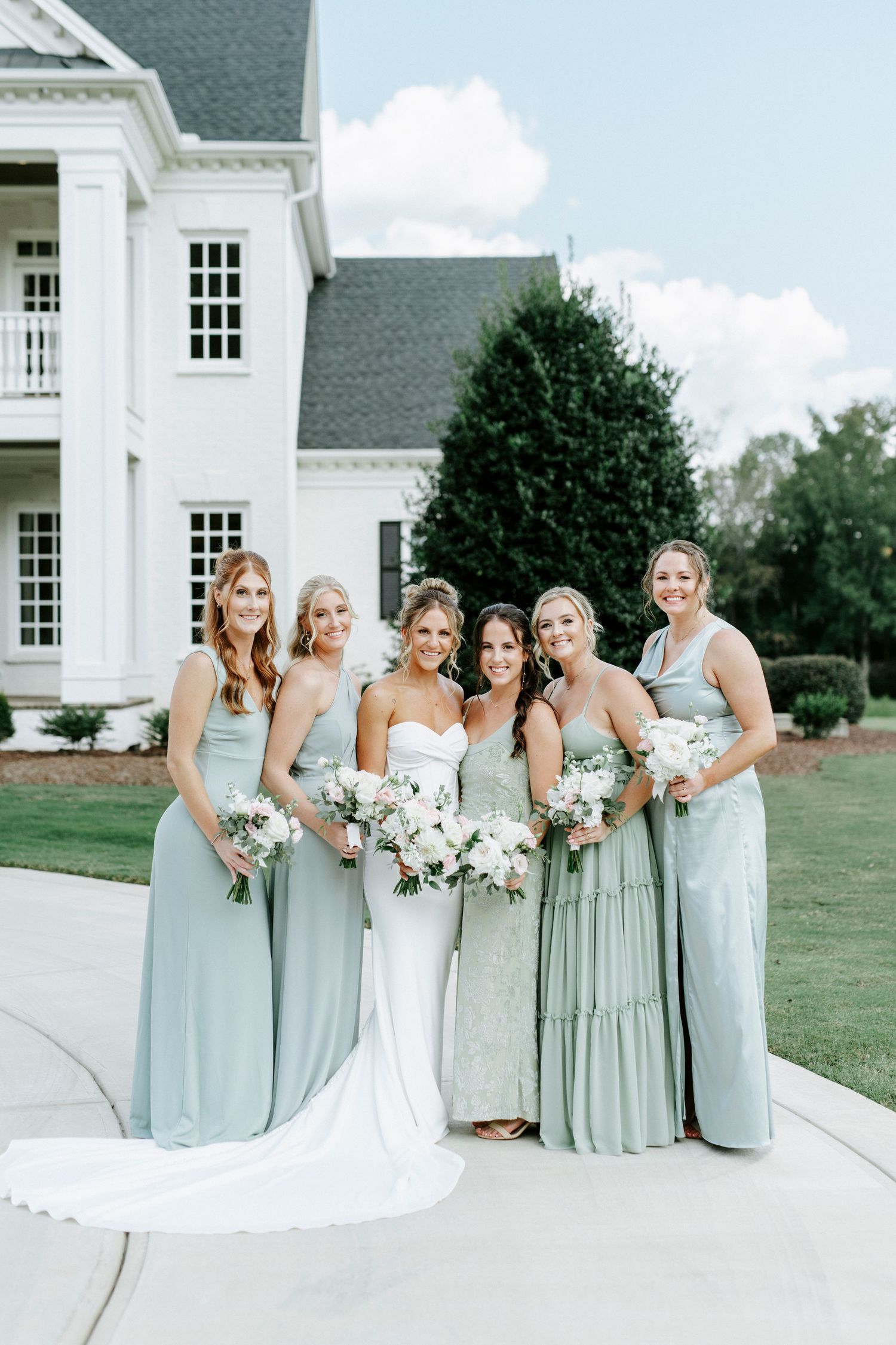 Wedding party wearing sage green bridesmaid dresses poses together on white stone patio near elegant mansion venue.