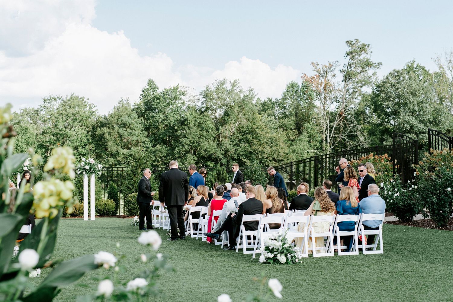 Wedding ceremony guests seated on white chairs in a lush outdoor garden setting during a summer celebration.