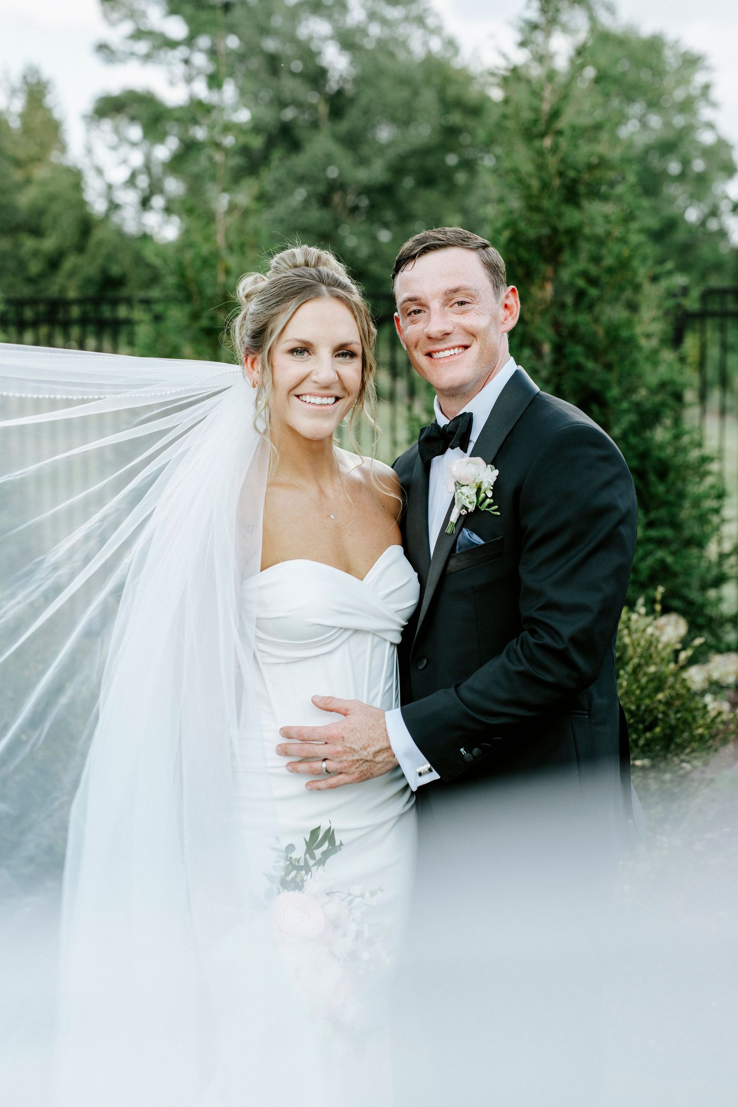 An elegant wedding portrait featuring a flowing veil and classic black tie attire.