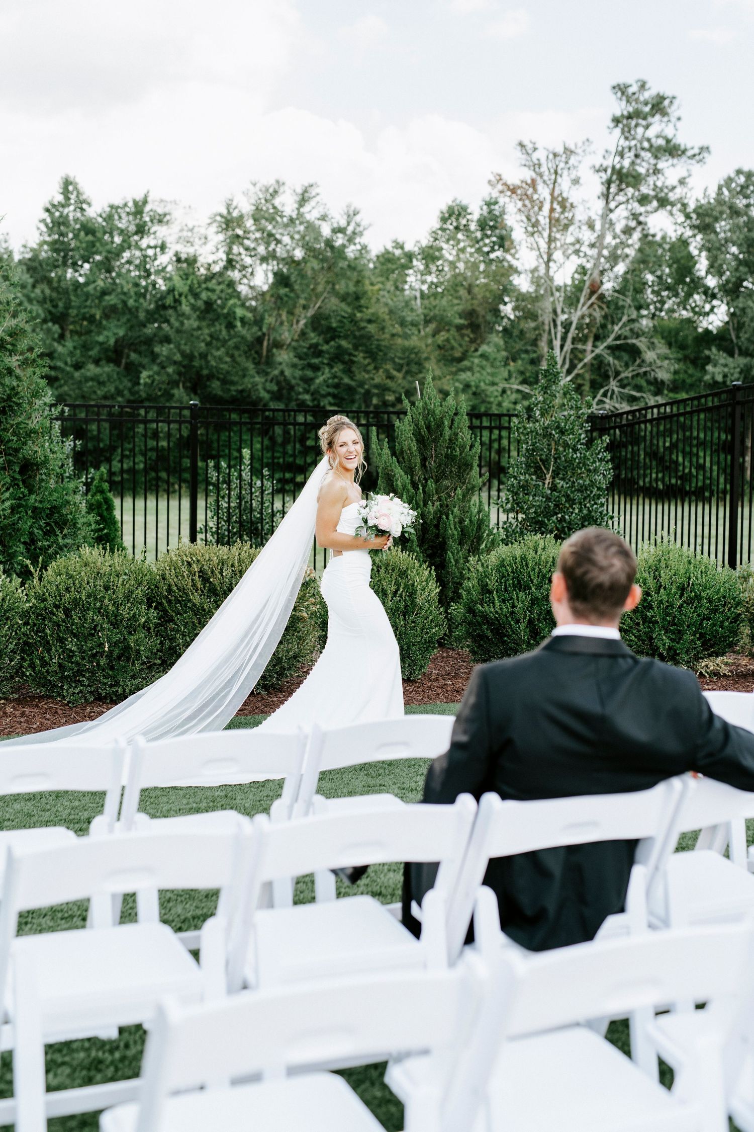 A bride in a flowing wedding dress and veil stands outdoors by a black iron fence while someone sits in white chairs.