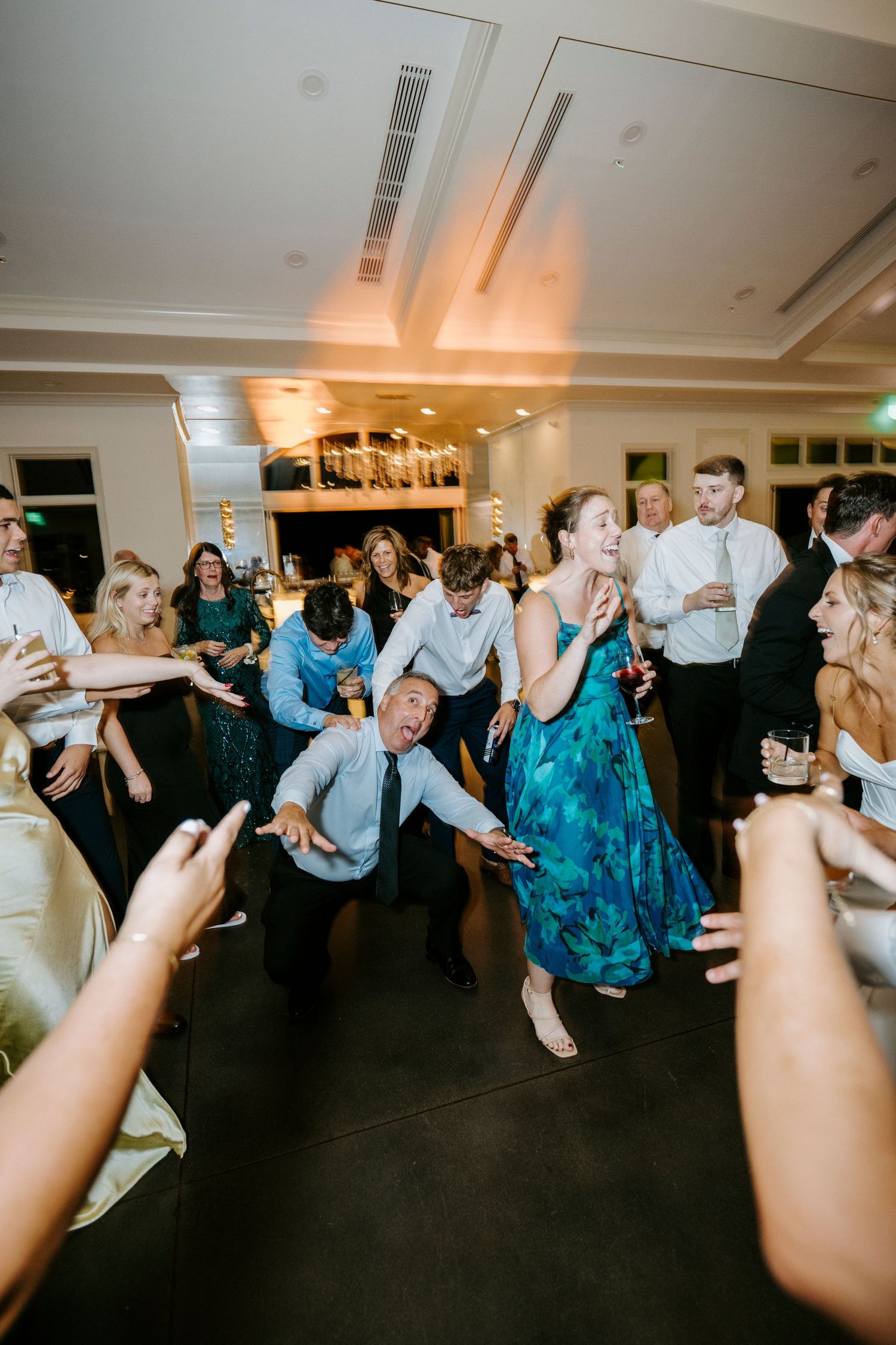 Wedding reception guests dance and celebrate on a dark dance floor under bright indoor lighting during a lively party.
