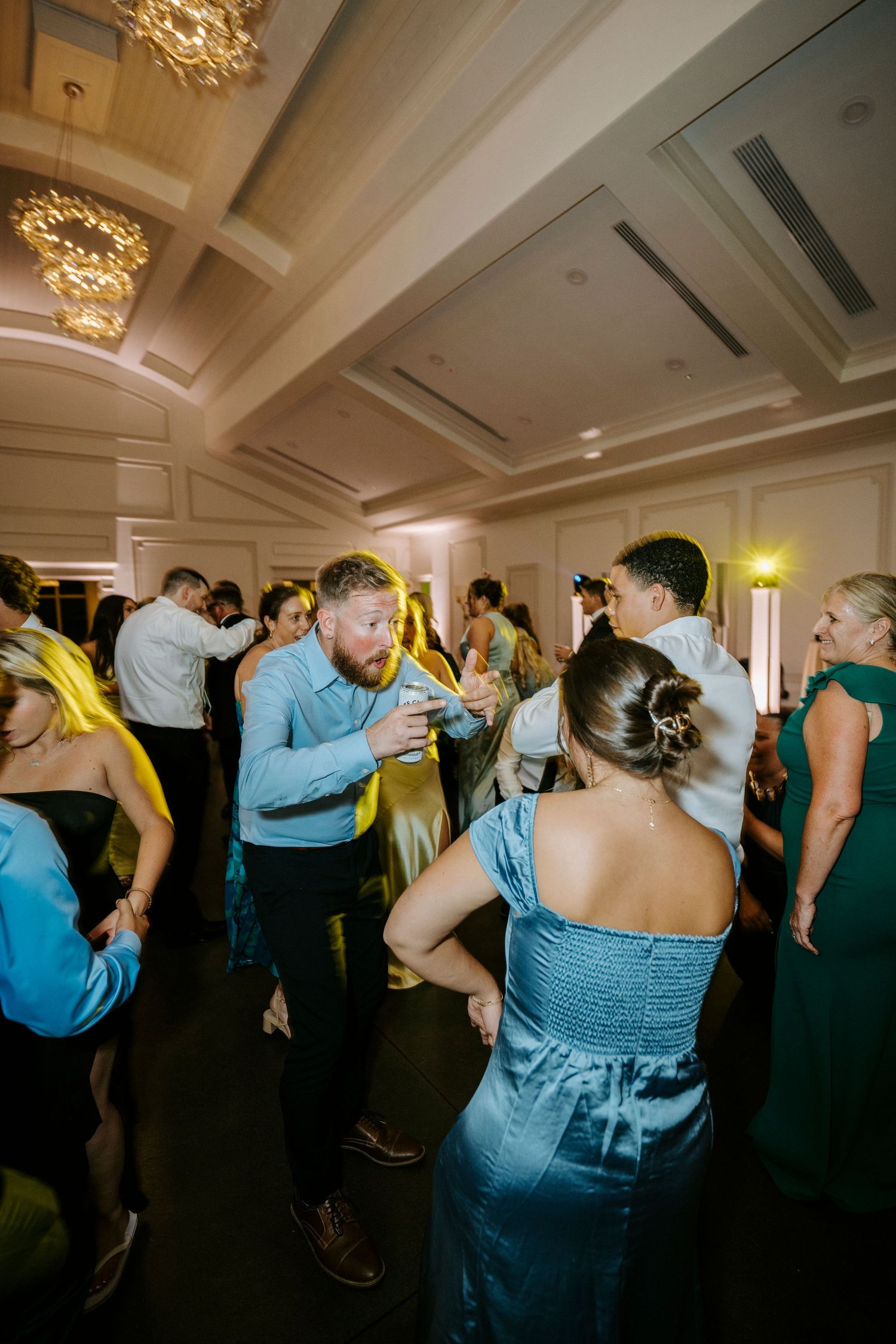 Wedding guests dance together on a lively dance floor under ornate crystal chandeliers at an elegant indoor reception.