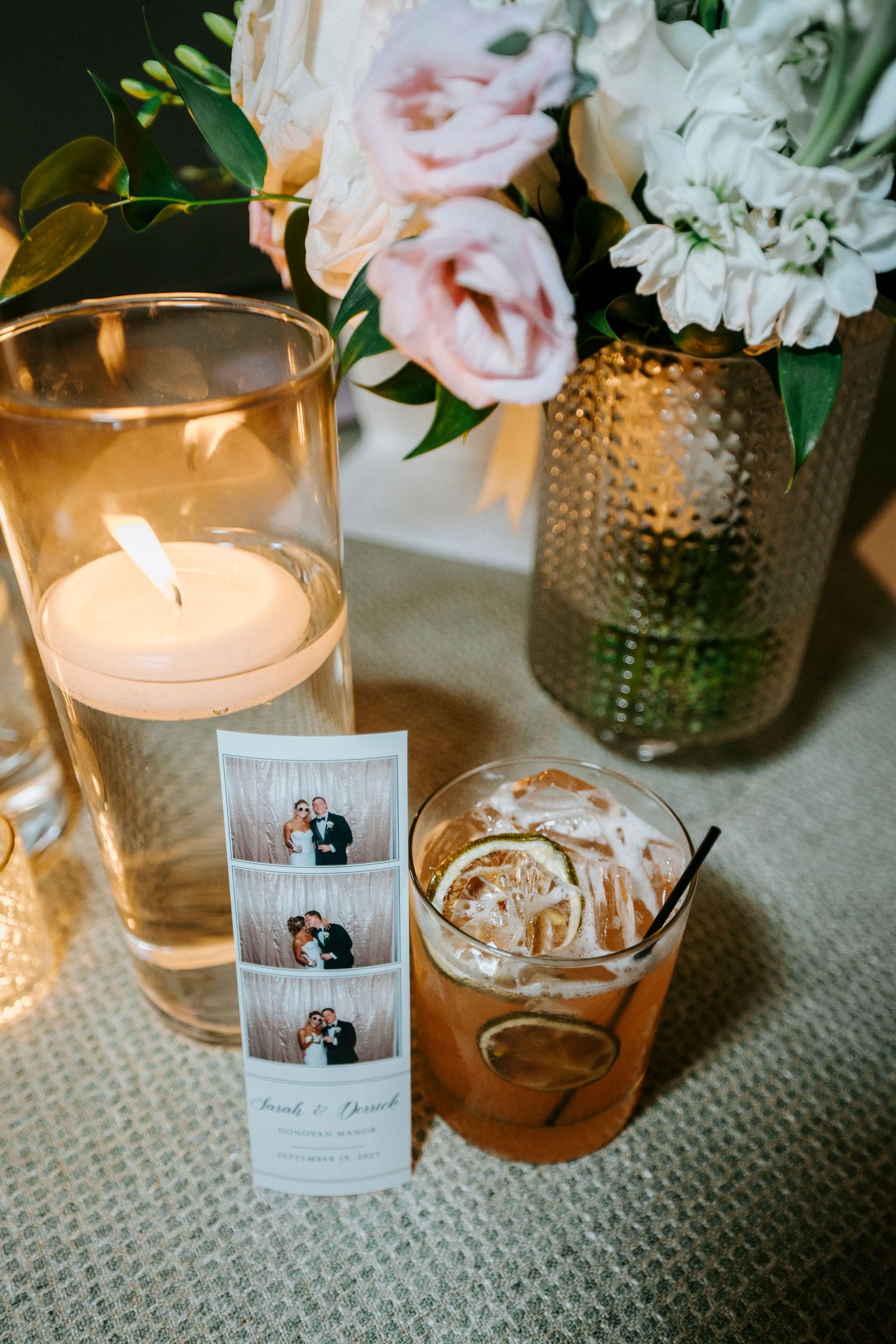 Wedding reception details with a glowing candle, photo strip, cocktail, and floral arrangements on a textured surface.