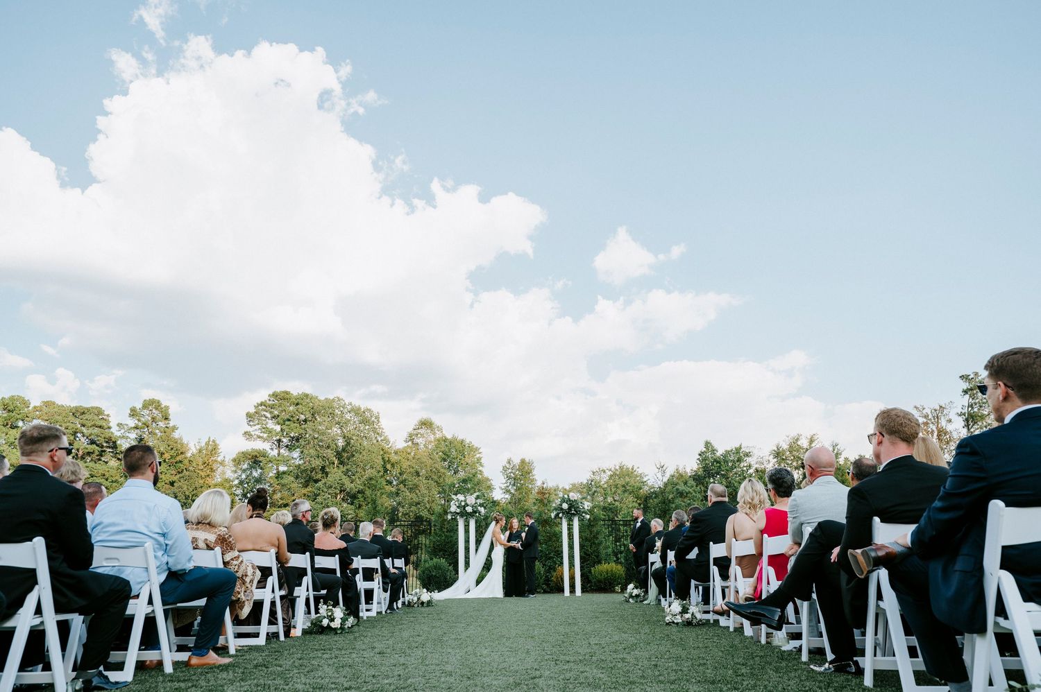Outdoor wedding ceremony setup with white chairs arranged in rows on green lawn under blue sky.