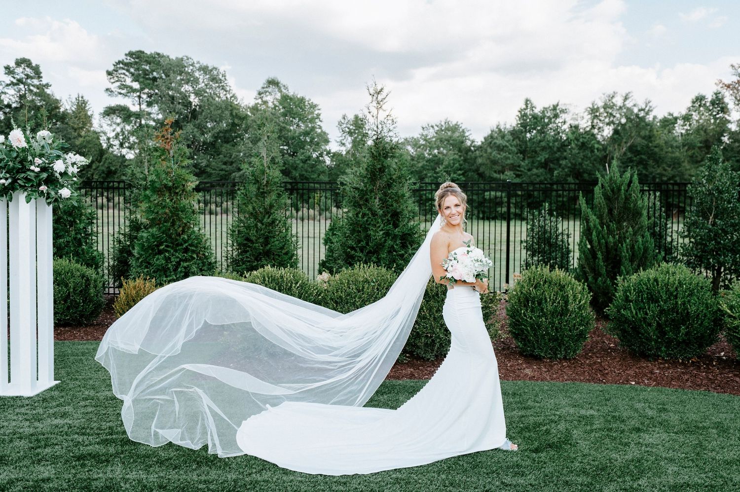 Flowing white wedding veil caught in breeze against lush garden backdrop creates ethereal outdoor portrait.